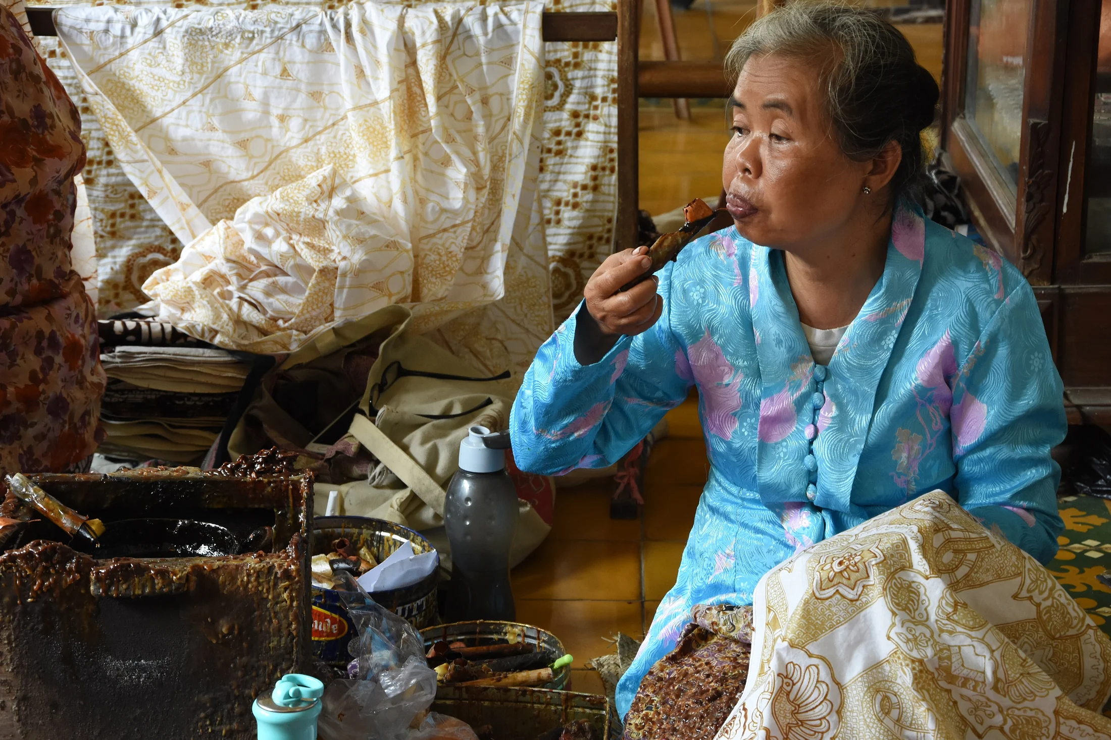 A woman in blue clothing sits beside batik fabrics and craft tools.